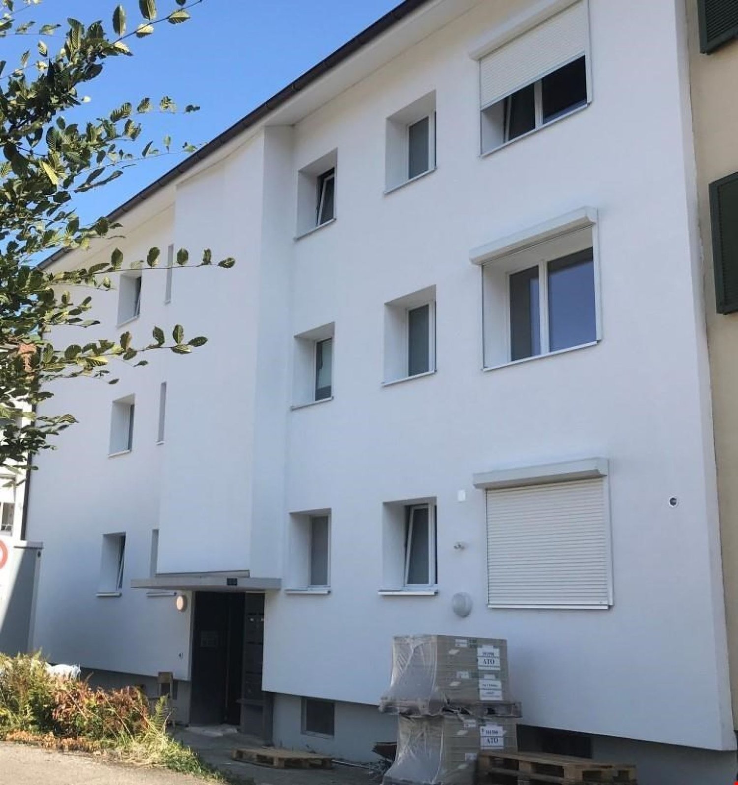 3 story white building, 4 windows per floor, entrance with door, small garden, and bushes, delivery packages near entrance