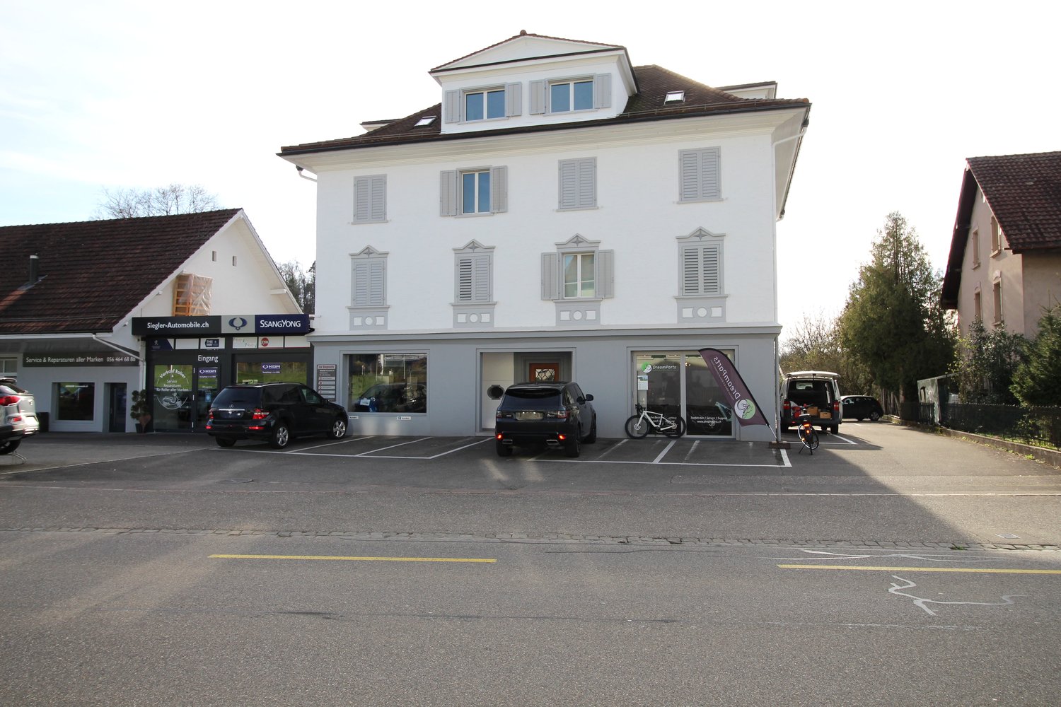 3 story house, white exterior, multiple windows, brown roof, parked cars in front, large paved parking area