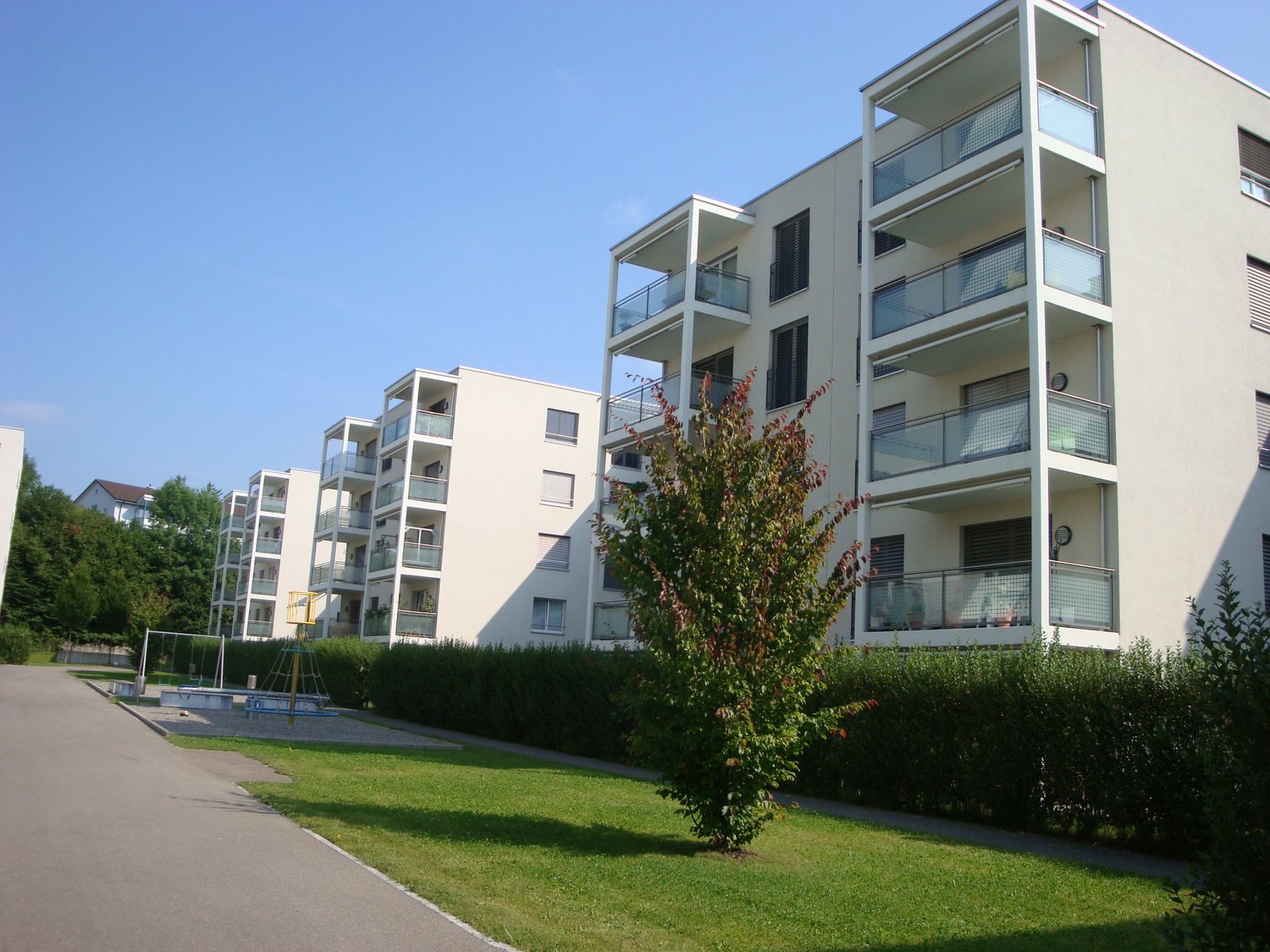Modern apartment building with multiple balconies, white exterior, greenery, large windows, outdoor playground, hedges, driveway