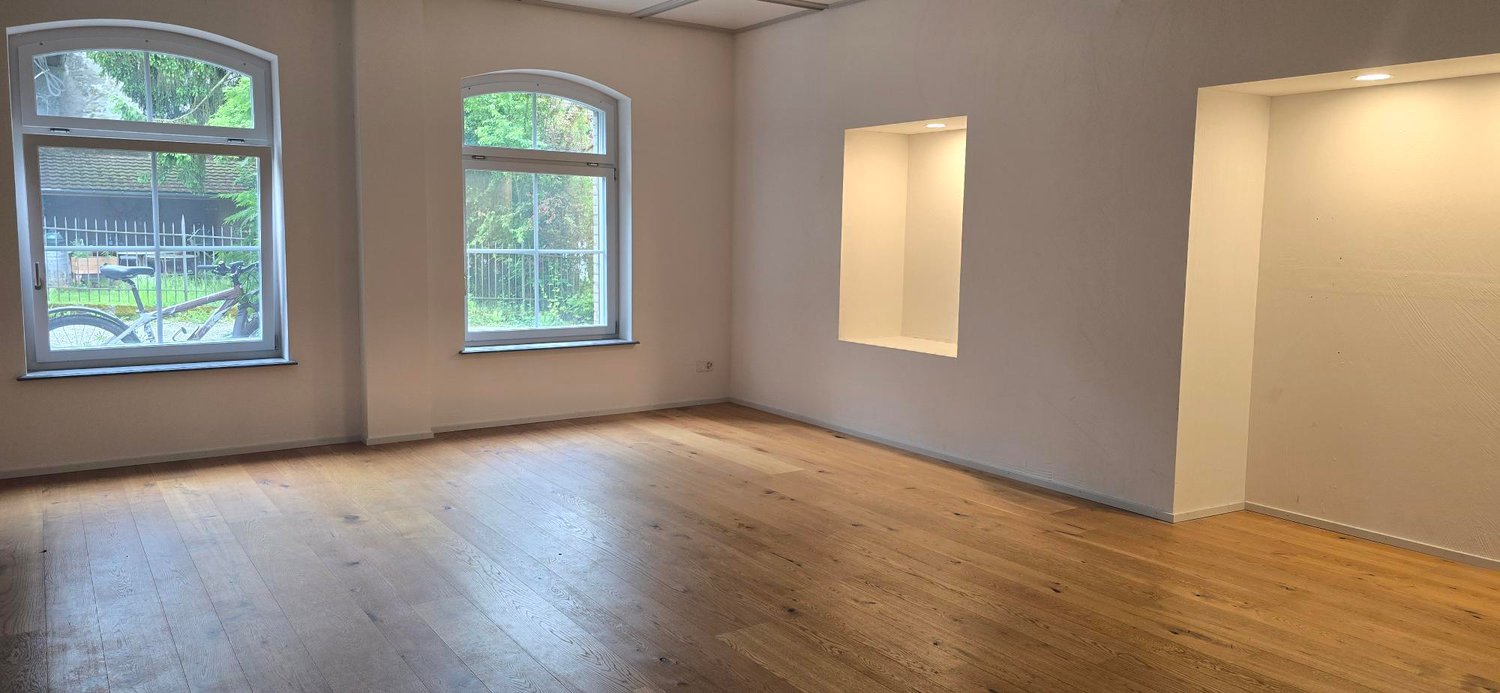 Large room with hardwood floors, arched windows overlooking a balcony or terrace, and neutral-colored walls.
