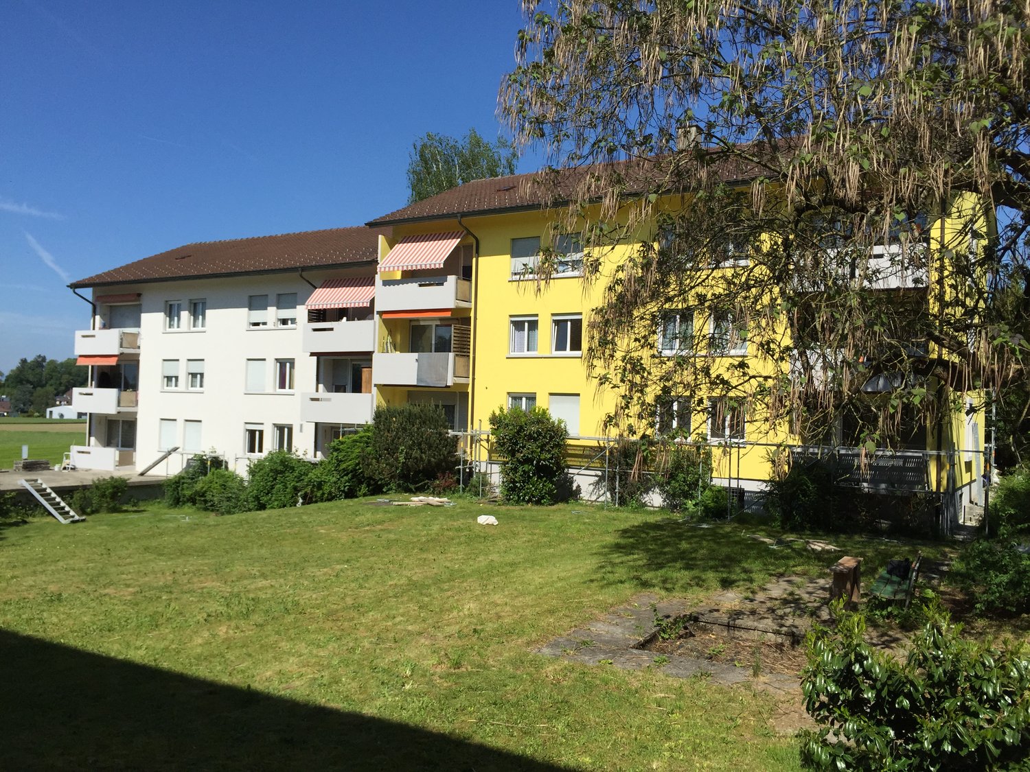 A two-story apartment building with a yellow facade, brown roof, white balconies, a staircase, and a green lawn in front.