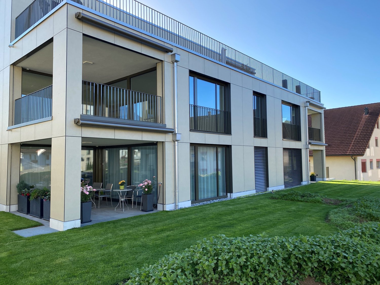 Modern 3 story apartment building, concrete exterior, glass windows, balconies, green lawn, potted plants