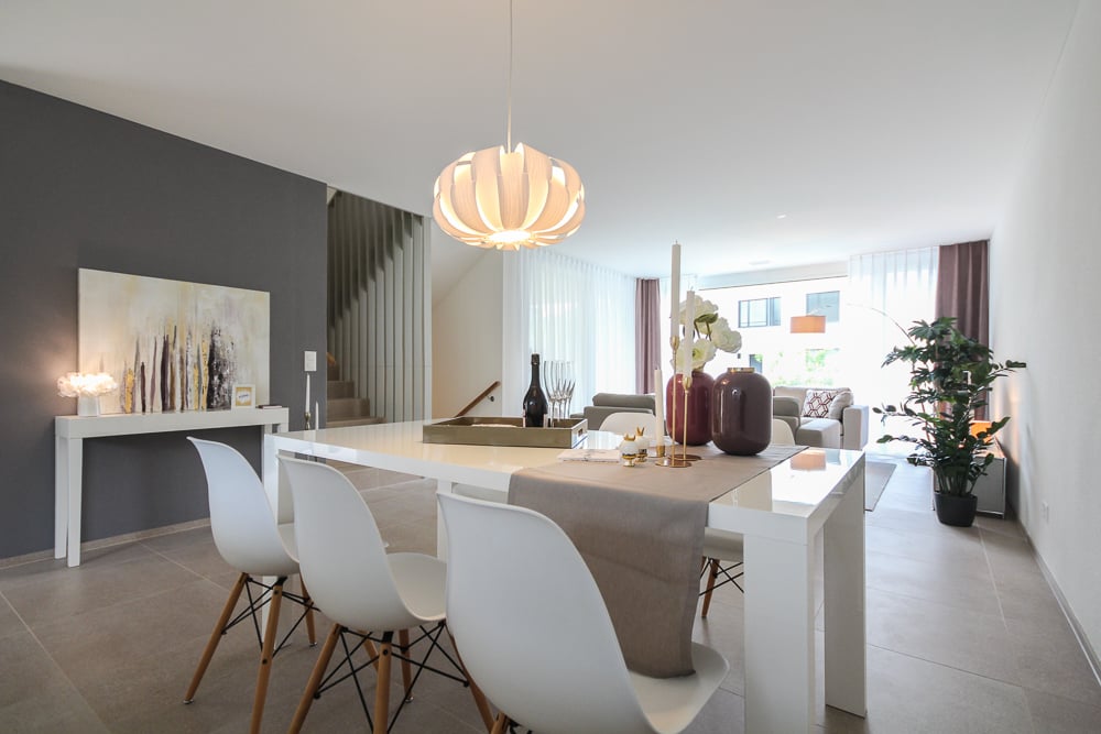 Spacious dining area with a modern white dining table and chairs, a decorative lighting fixture, and a plant in the corner. The room has gray walls and a tiled floor, creating a sleek and minimalist design.