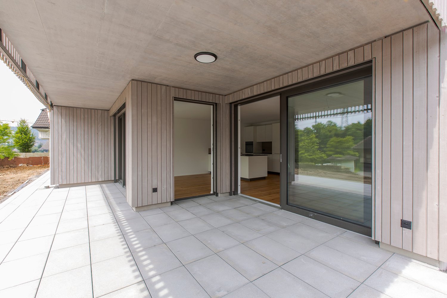 Spacious covered balcony or terrace with large sliding glass doors, wooden paneled walls, tiled flooring, and recessed lighting in the ceiling.