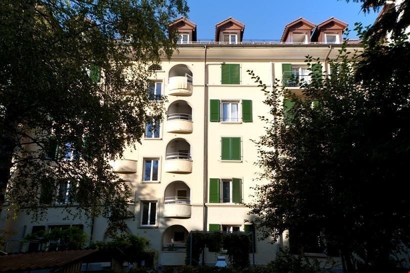5-story apartment building, cream-colored exterior, several balconies, green shutters, rooftops with red tiles, large trees in front