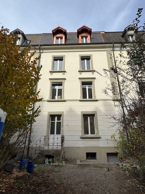 4-story building, multiple windows, gray roof, located in the street, surrounded by trees