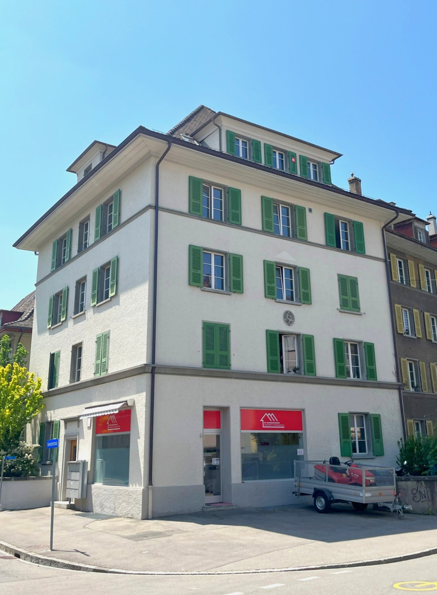 3 story house, white wall, green shutters, 2 shops on the ground floor, small garden in front of the building
