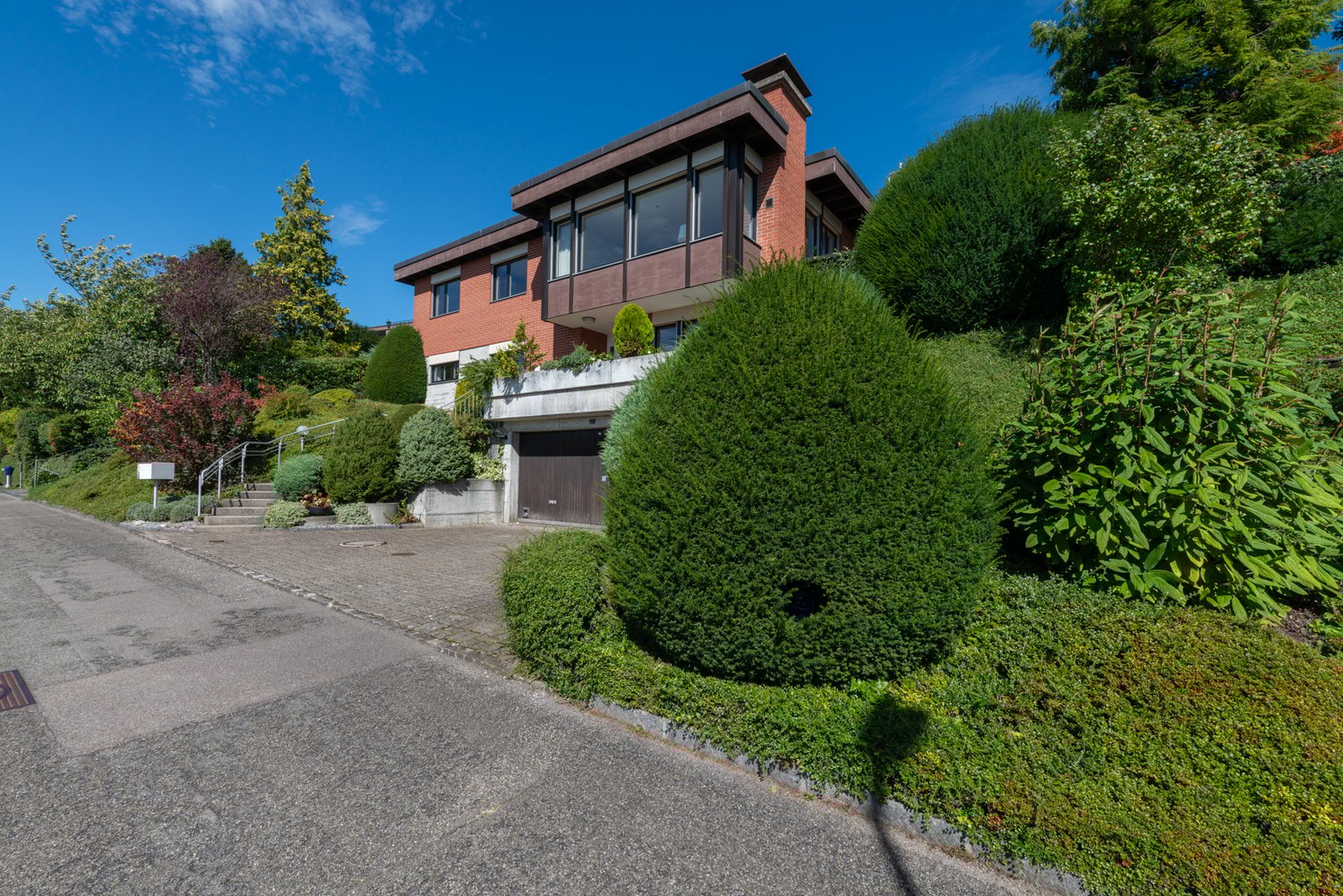Modern house, elevated on slope, red brick exterior, garage, large windows, multiple levels, landscaped garden with shrubs