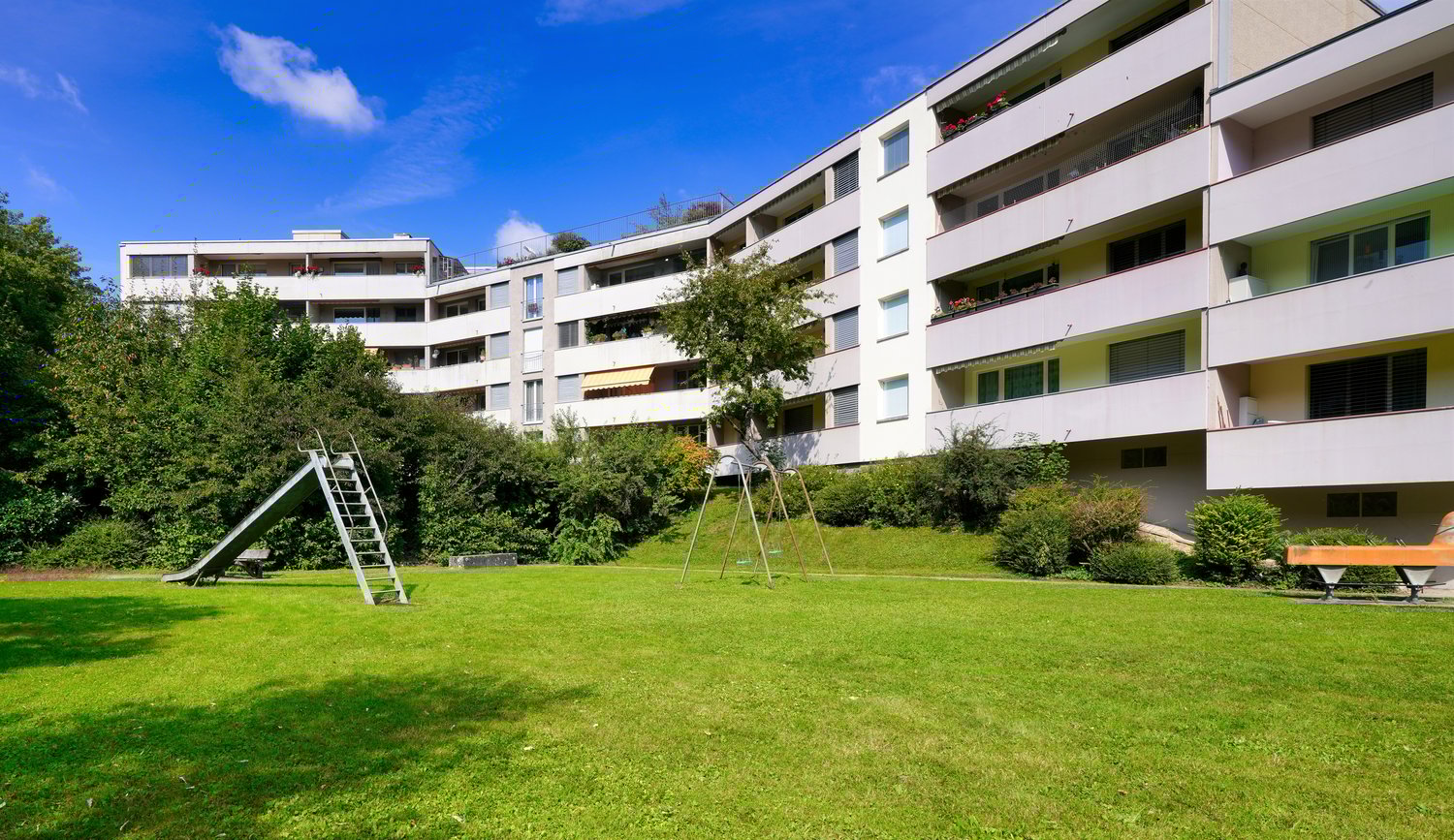 green lawn, trees, bushes, playground with slide and swings, building with balconies in the background
