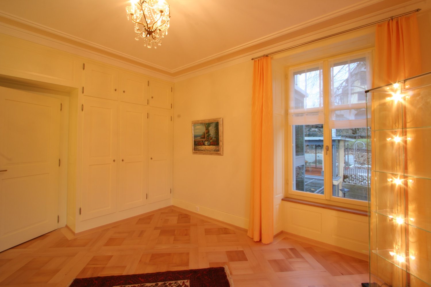 wooden floor, chandelier, white wardrobe, yellow walls, picture frame, orange curtains, large window, glass cabinet