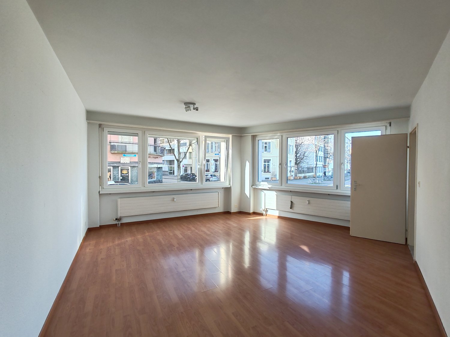 Empty room, wooden floor, 4 windows, white walls, 2 radiators, an entrance door