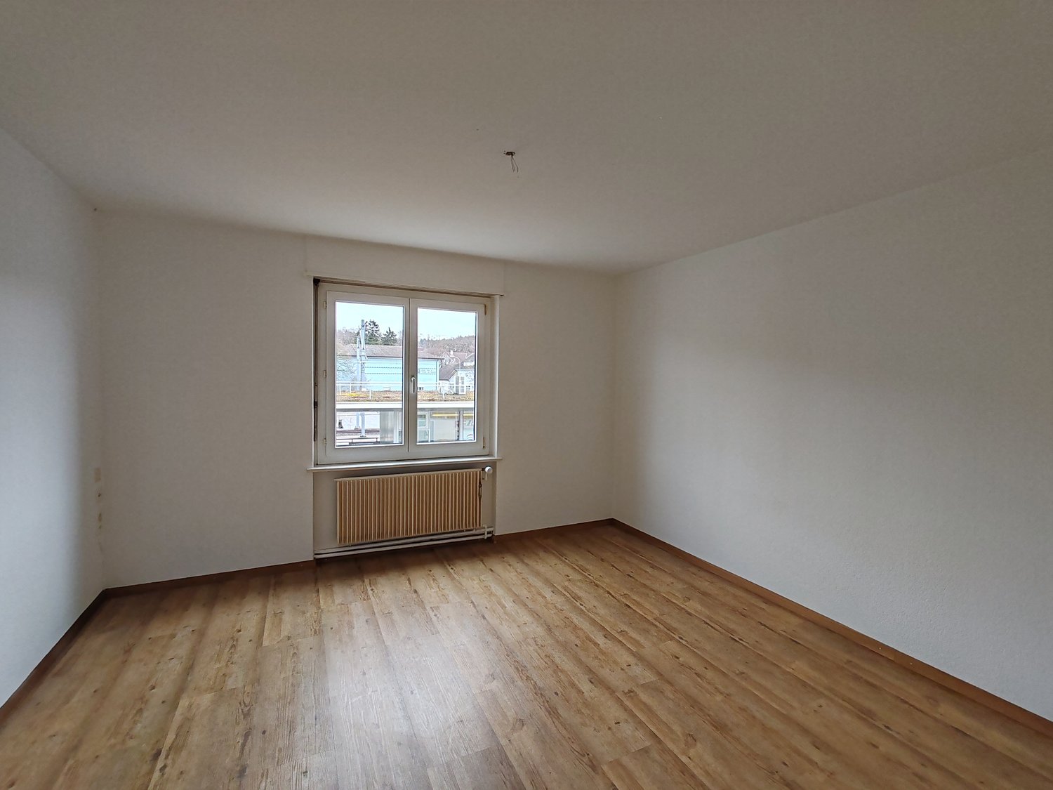 An empty room with white walls, wooden floor, a window, and a radiator.
