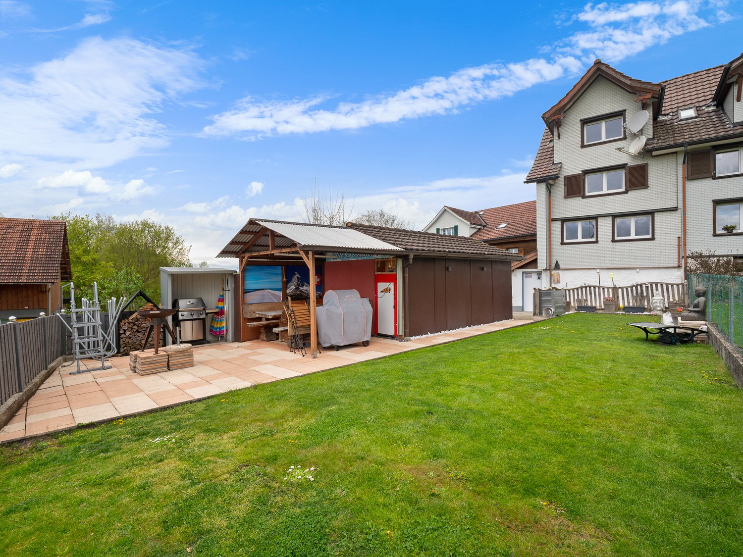 Two-story residential building with a tiled roof, surrounded by a grassy yard and a paved patio area. The yard features a wooden shed or storage structure, a barbecue grill, and other outdoor furniture. There is also a garage or carport visible on the pro