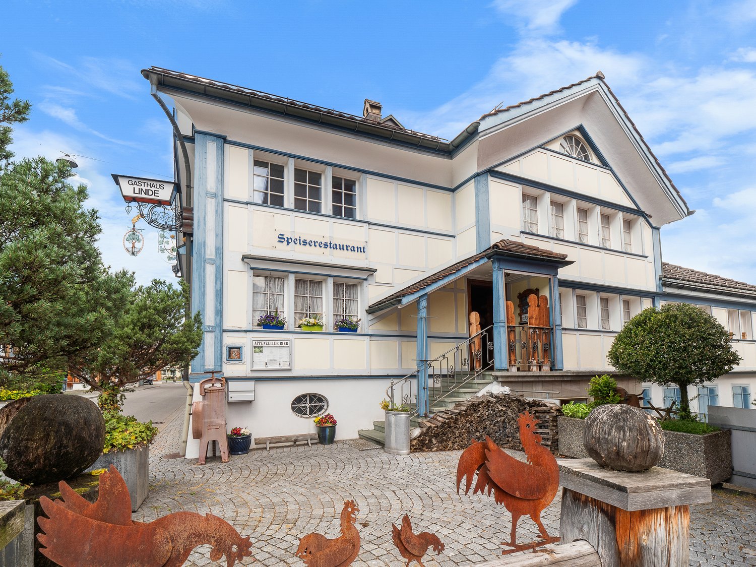 Two-story building with a traditional German architectural style, featuring a restaurant called 'Speiserestaurant' on the ground floor. The building has a tiled roof, large windows, and decorative elements. There are some rustic metal sculptures of animal