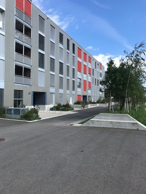 Gray apartment building with red shutters, grey pavement, several parking spaces, trees and plants