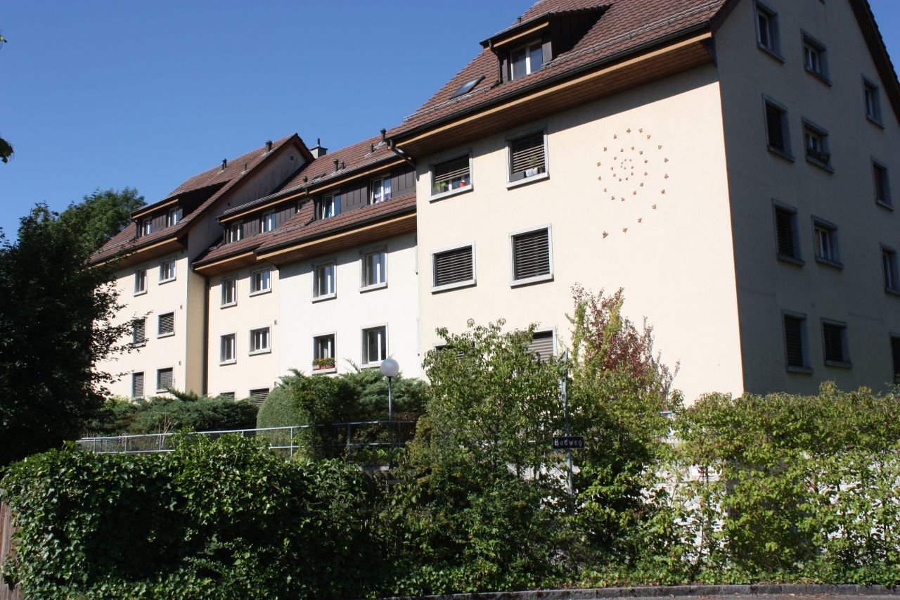 large white building with several floors, multiple balconies, brown roof, greenery in front