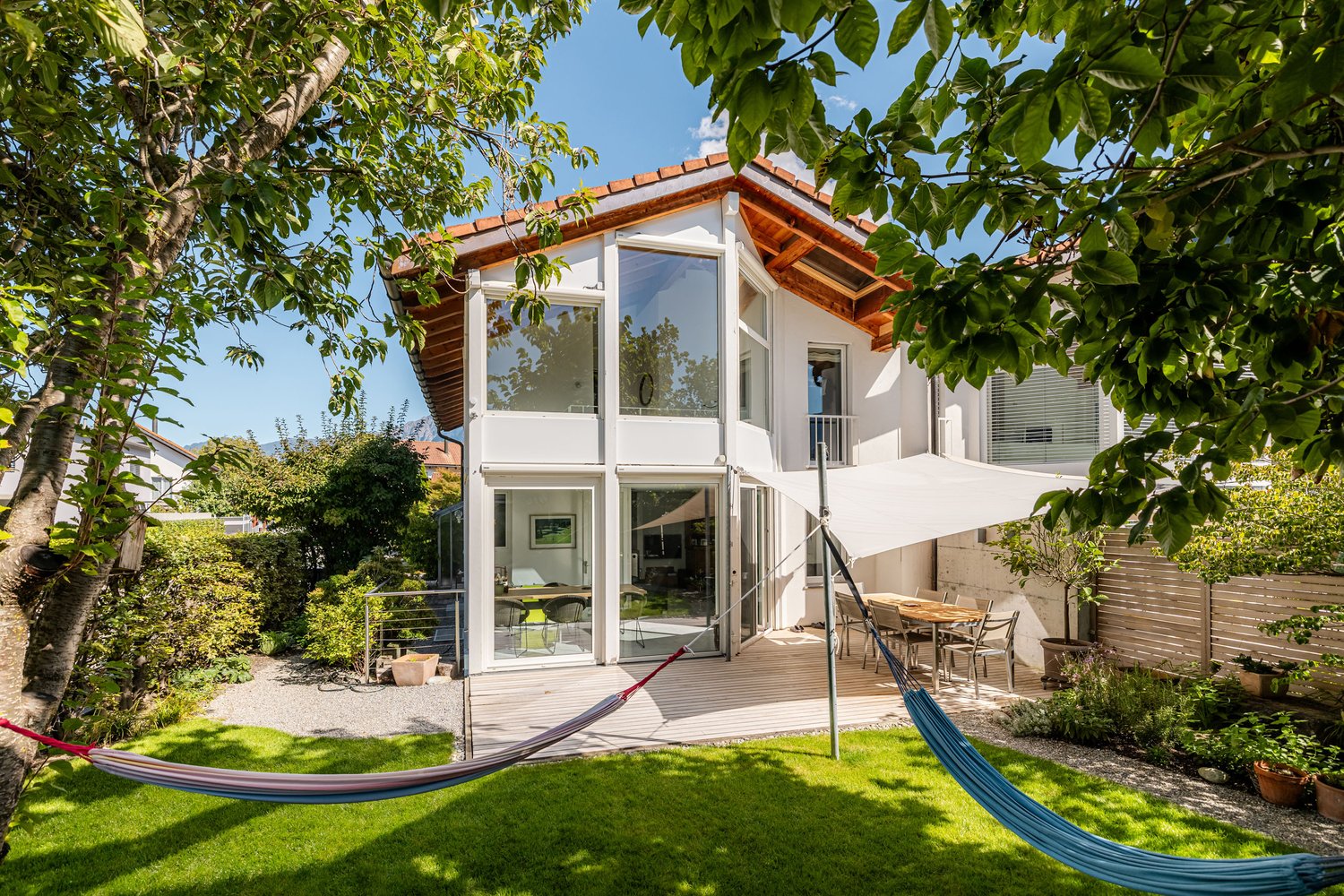 white house, large glass windows, wooden deck, outdoor dining area, hammocks, garden with trees