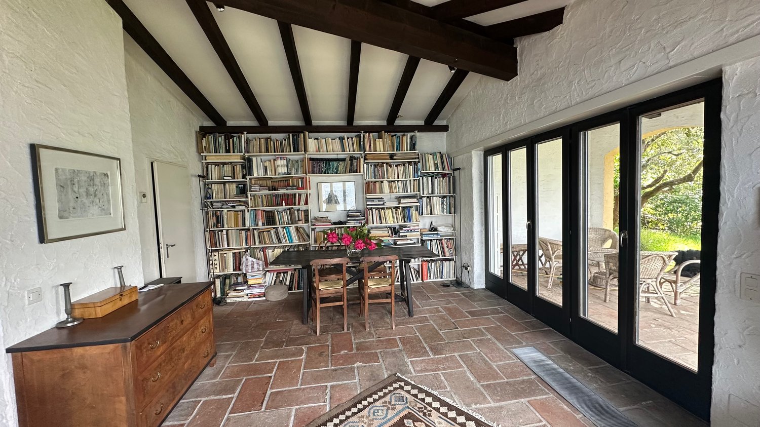 Dining room with wooden table, chairs, bookshelves, brick floor, sliding glass doors, picture frame, flowers, rug, wooden cabinet, outdoor seating