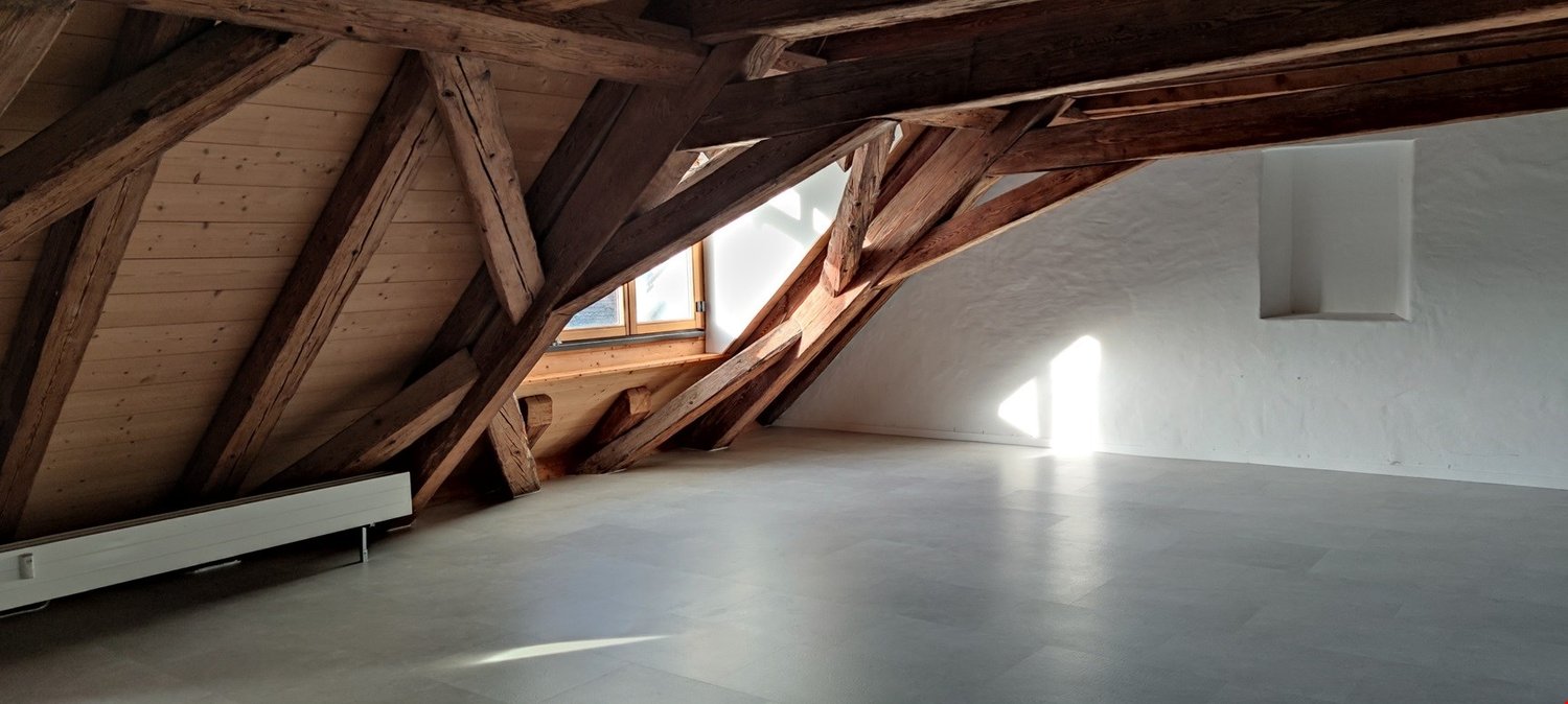 Spacious attic with exposed wooden beams and trusses, large windows providing natural light, concrete floor, and potential for conversion or storage space.