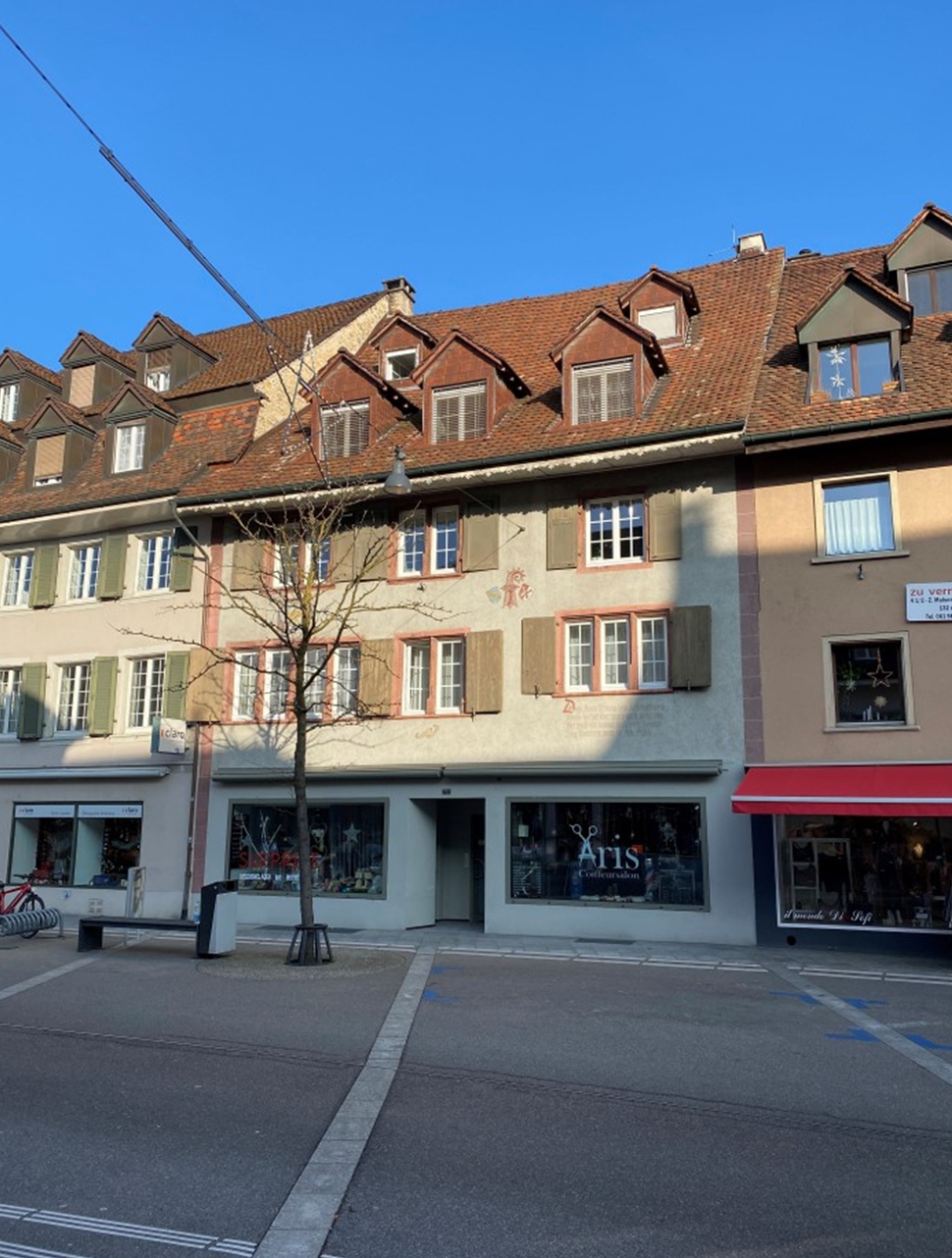 Multi-story building with red tiled roofs, dormer windows, and a mix of residential and commercial spaces. The building has a traditional European architectural style with a cobblestone street in front.