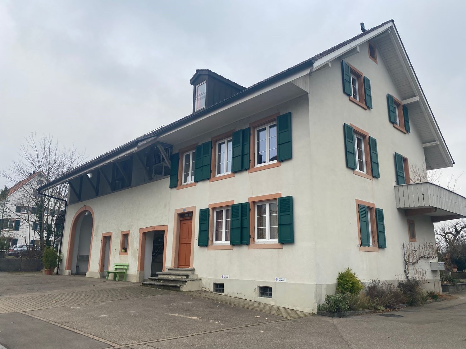 A two-story building with a traditional European architectural style, featuring a light-colored exterior, green shutters, and a slanted roof. The building has several windows and a covered entrance area.