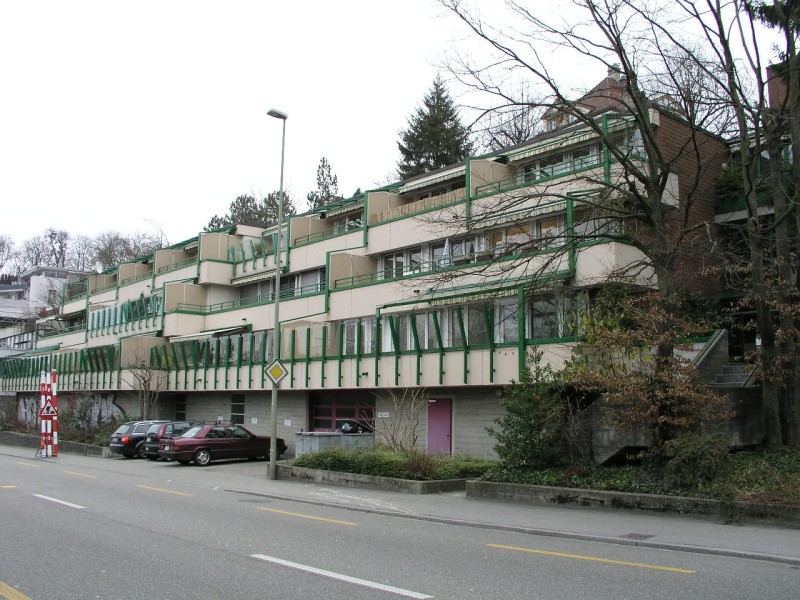 Multi-story apartment building with balconies, brick and concrete construction, surrounded by trees and vegetation