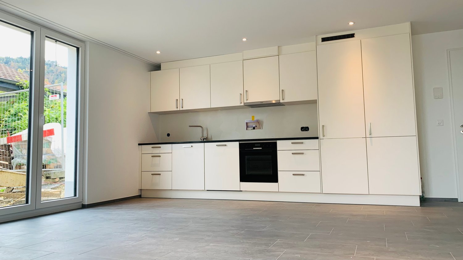 Spacious modern kitchen with white cabinets, black countertops, built-in oven, and dishwasher. Large windows provide natural light and a view of the surrounding area.