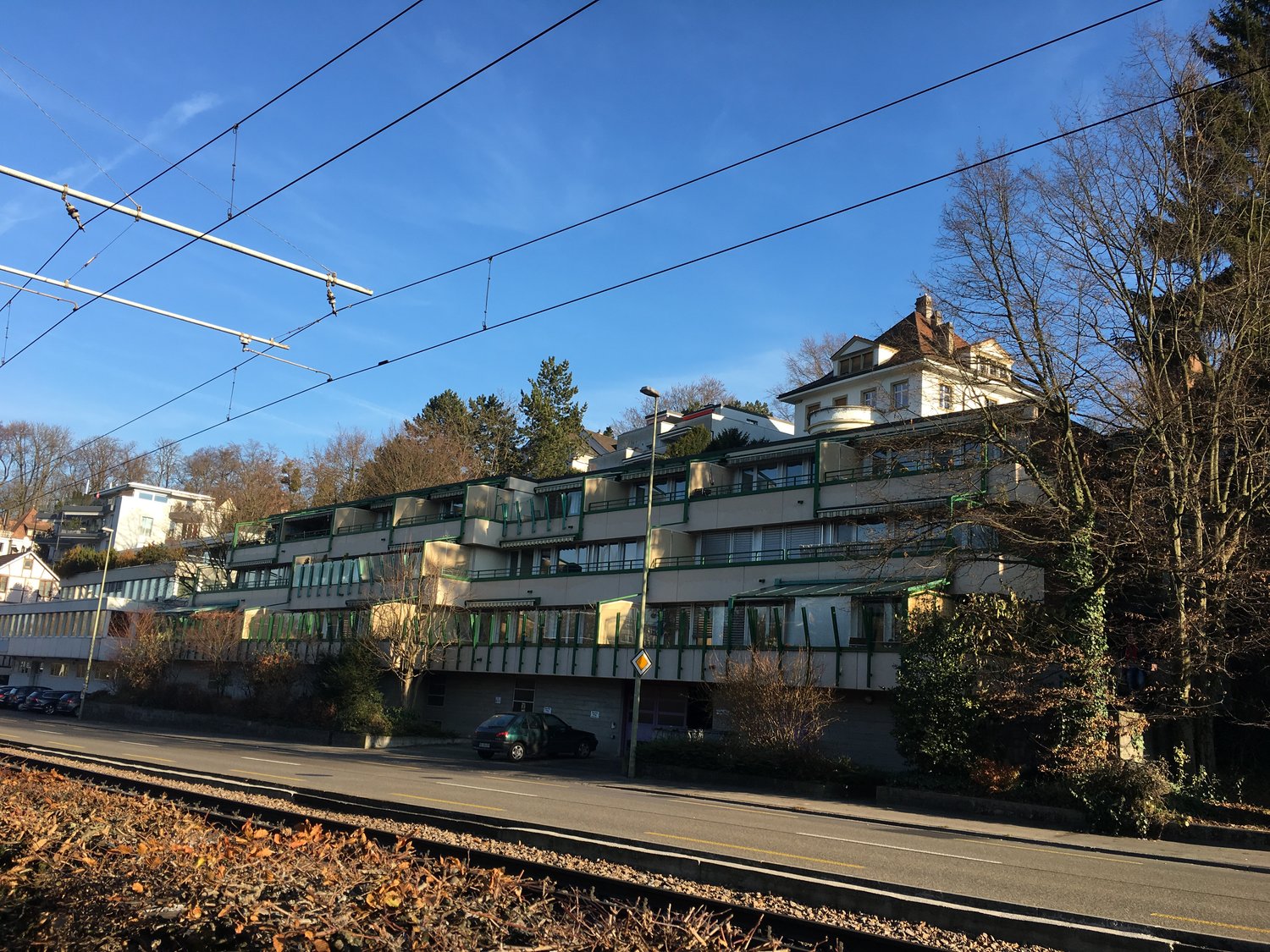 Large building, multiple floors, balconies, green windows, parked car