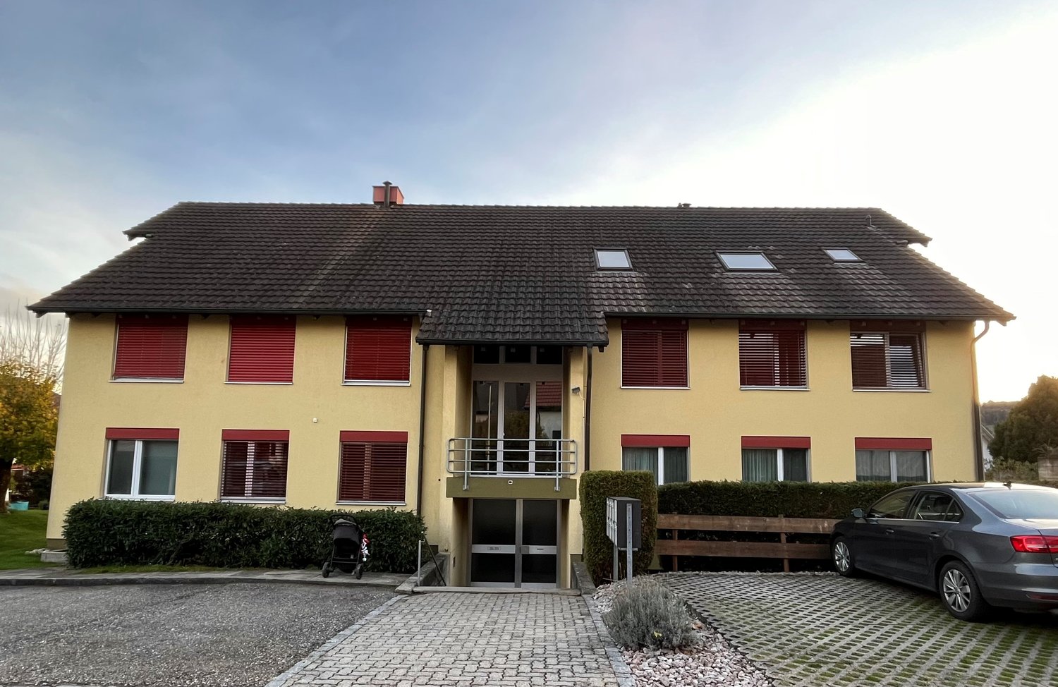 2-story yellow house, black roof, red shutters, glass doors, small plants, parking lot, car parked, fence