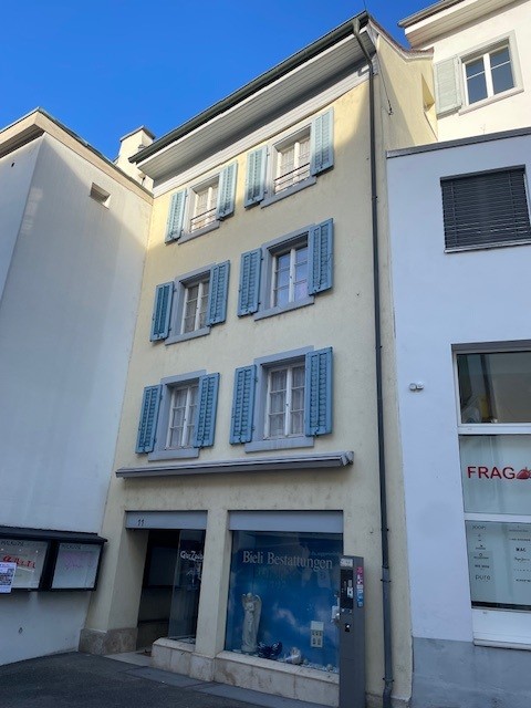An apartment building with several windows and blue shutters, a yellow painted wall, a black door at the entrance and a display glass in the bottom floor.