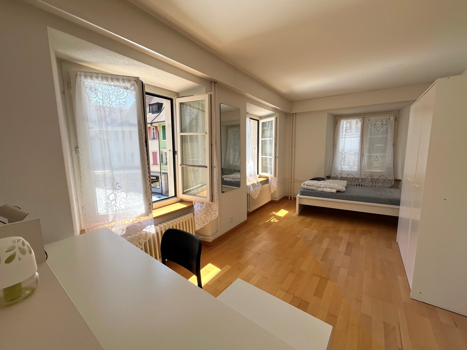 wooden floor, white walls, open windows, bed, desk, chair, white curtains, radiator, small decorative object on desk