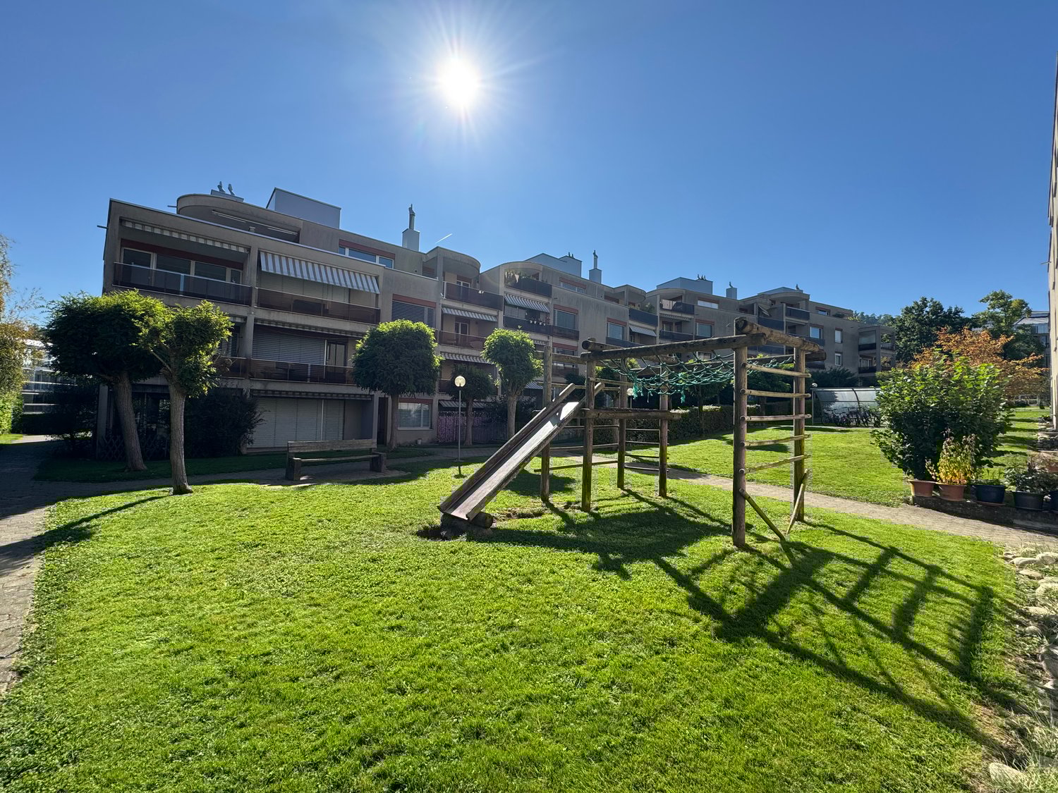 Playground with a slide and a bench, surrounded by green grass and trees, with a walkway leading to the building