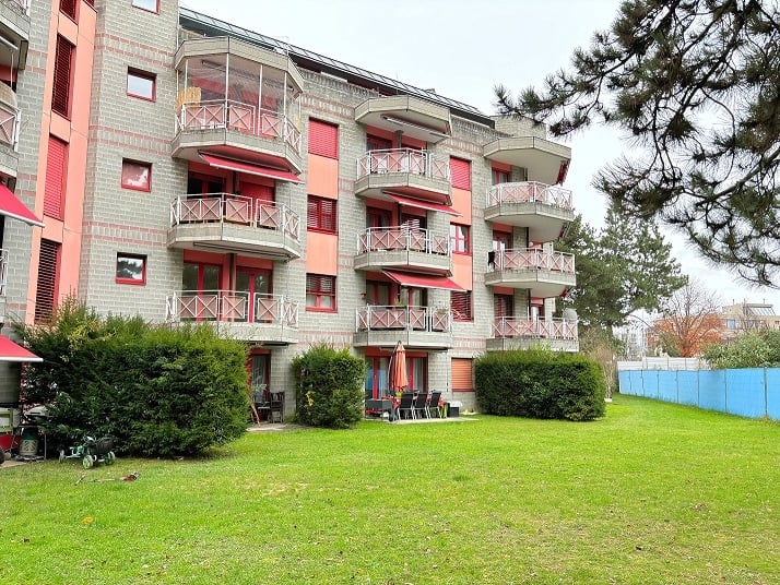 The building has several balconies, brick walls, awnings, and windows. Outside, there are tables and chairs with an umbrella on the grass lawn, and some plants and trees.