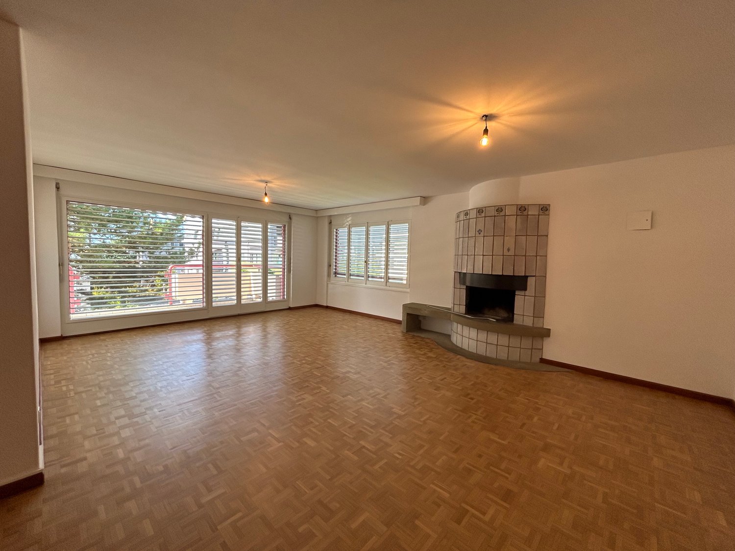 wooden parquet flooring, fireplace on wall, glass windows, sliding glass doors, white walls, two ceiling lamps