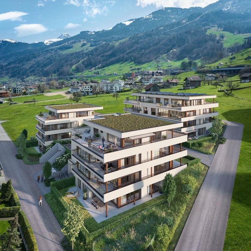Multi-story apartment building with balconies, surrounded by green hills and mountains in a scenic alpine landscape