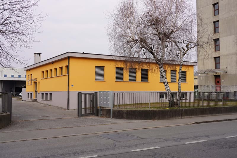 A single story yellow building with gray trims. A gate leads to the parking lot with trees in the center. Several windows and a chimney are visible on the yellow wall. A grey building is on the right side.