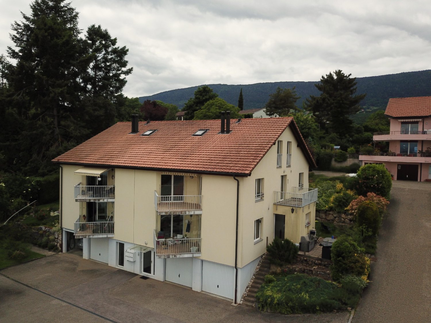 A multi-story apartment building with a red tile roof, balconies on each floor, and surrounded by trees and greenery. The building appears to be situated in a hilly, forested area with mountains in the background.