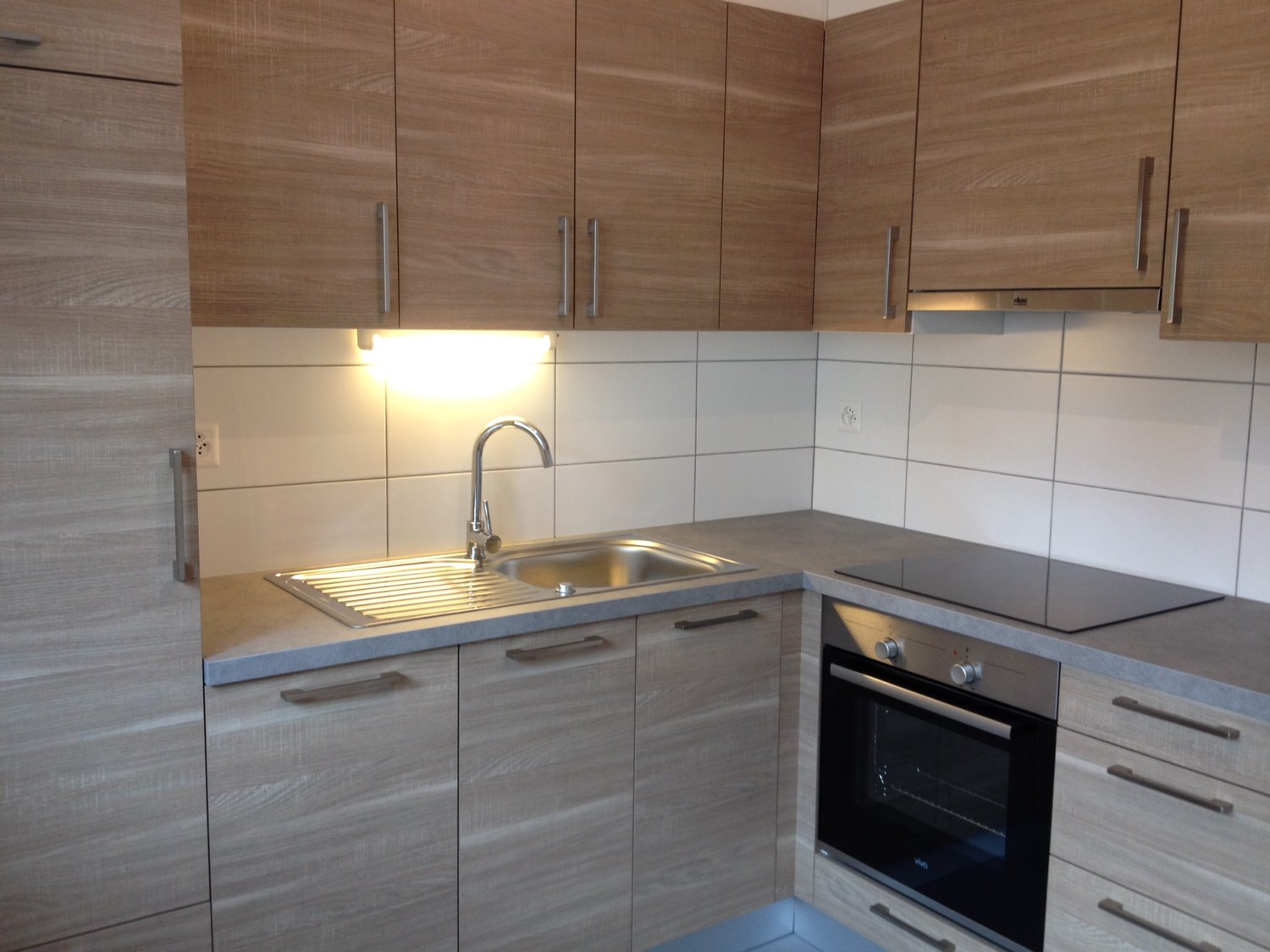 wooden cabinets, white tiled walls, stainless steel sink, black electric oven, countertop, bright lighting