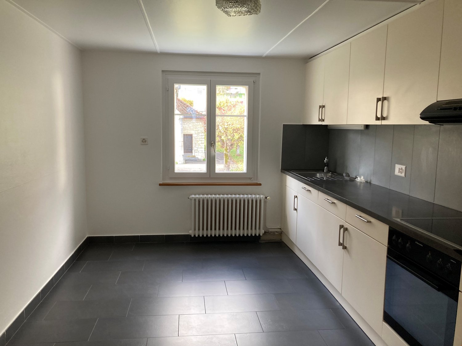 The image shows a modern kitchen with white cabinets, black countertops, and a tiled floor. There is a window overlooking a garden or outdoor area. The kitchen appears to be equipped with a stove and oven.