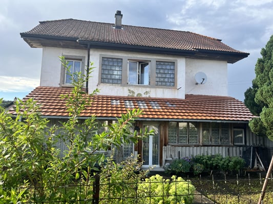 Two-story house with a tiled roof, white exterior walls, and several windows. There is a balcony or terrace visible on the upper level. The house is surrounded by trees and greenery.