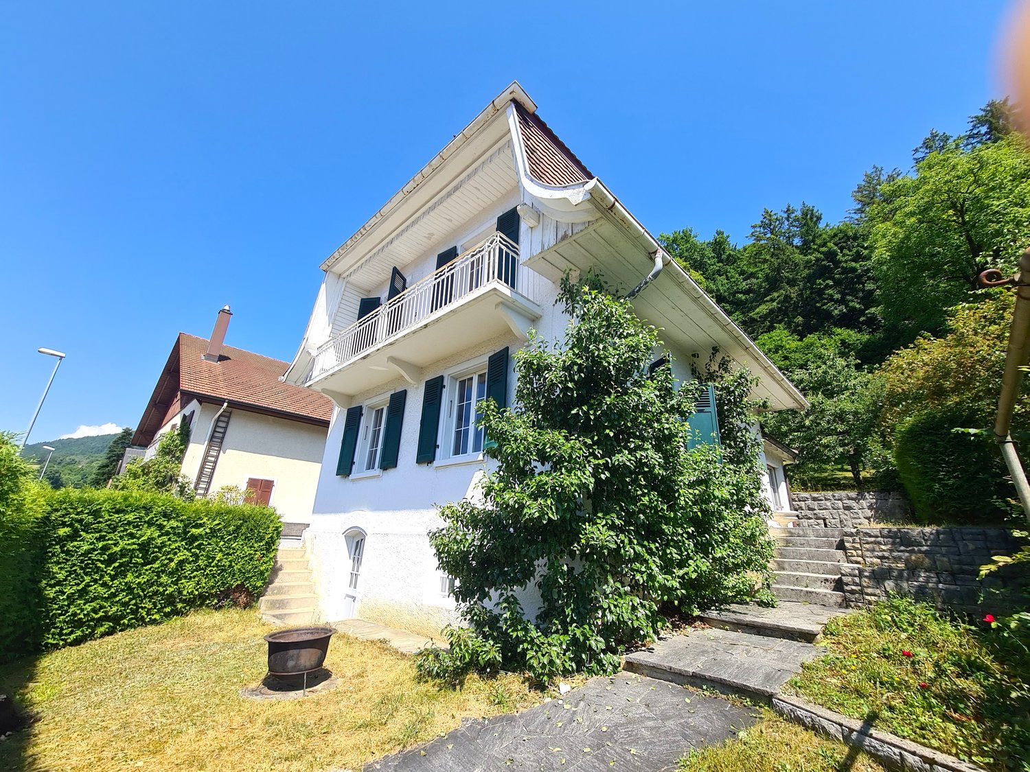 2-story house with white exterior, green shutters, balcony, surrounded by lush greenery and trees
