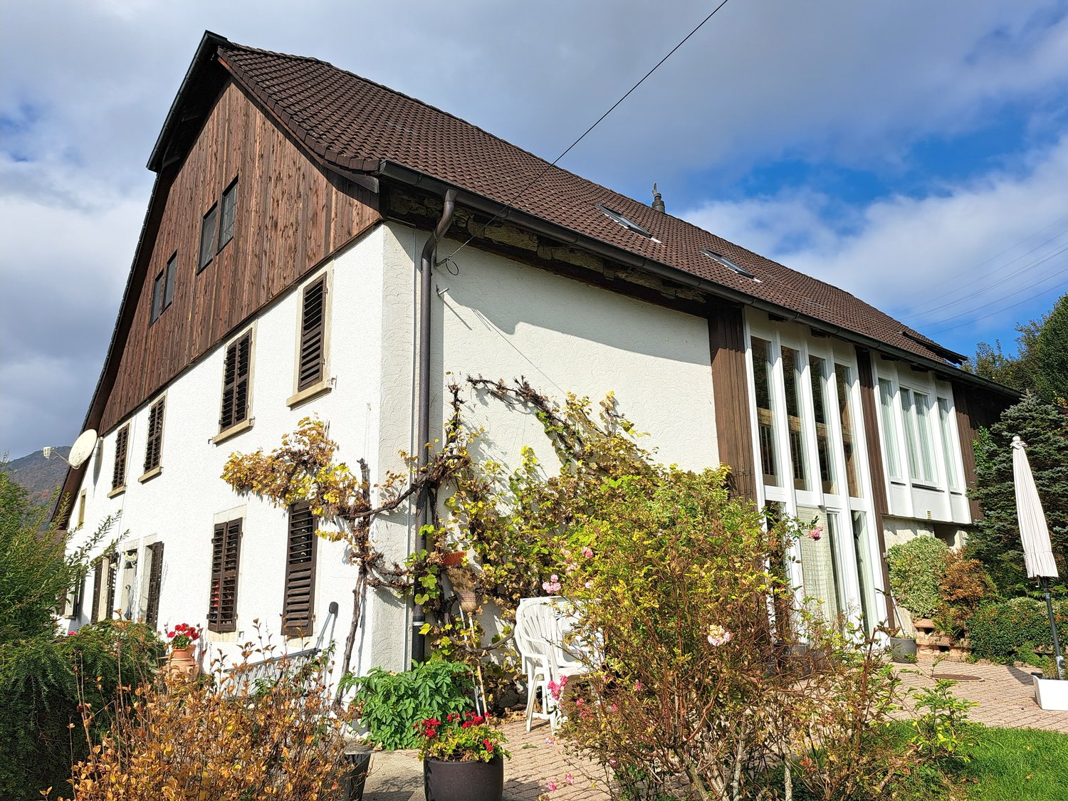 two-story house, brown roof, white wall, several windows, multiple potted plants, white lawn umbrella, greenery
