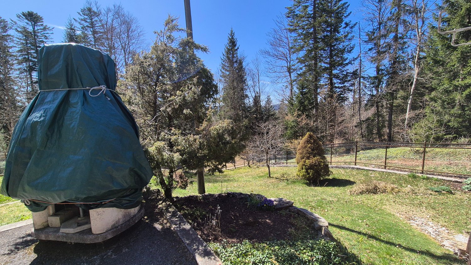 The image shows an outdoor area with a covered structure, likely a patio or gazebo, surrounded by trees and greenery. There is a grassy area with some landscaping features like shrubs and a small mound. The overall scene suggests a well-maintained, natura