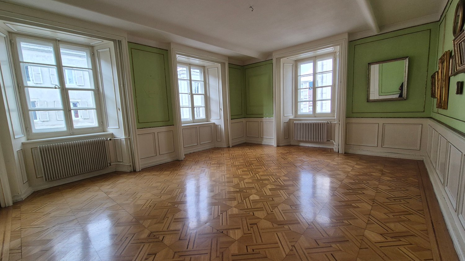room with wooden floor, green walls, 3 windows, radiators on both sides, mirror on right wall, pictures hanging on the wall