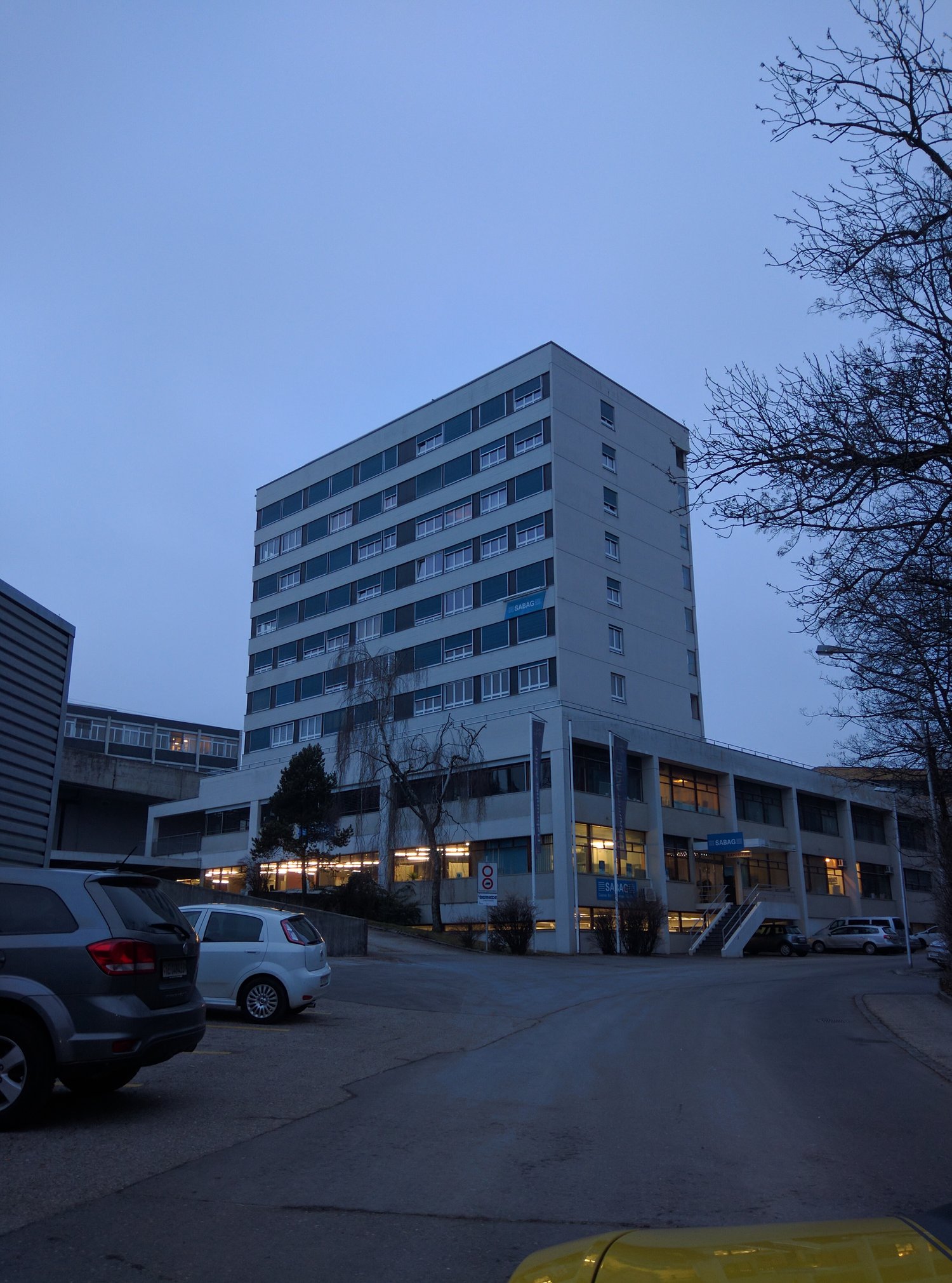 High rise building with multiple windows, glass doors, sign reading 'SABAD', cars parked on both sides of the street