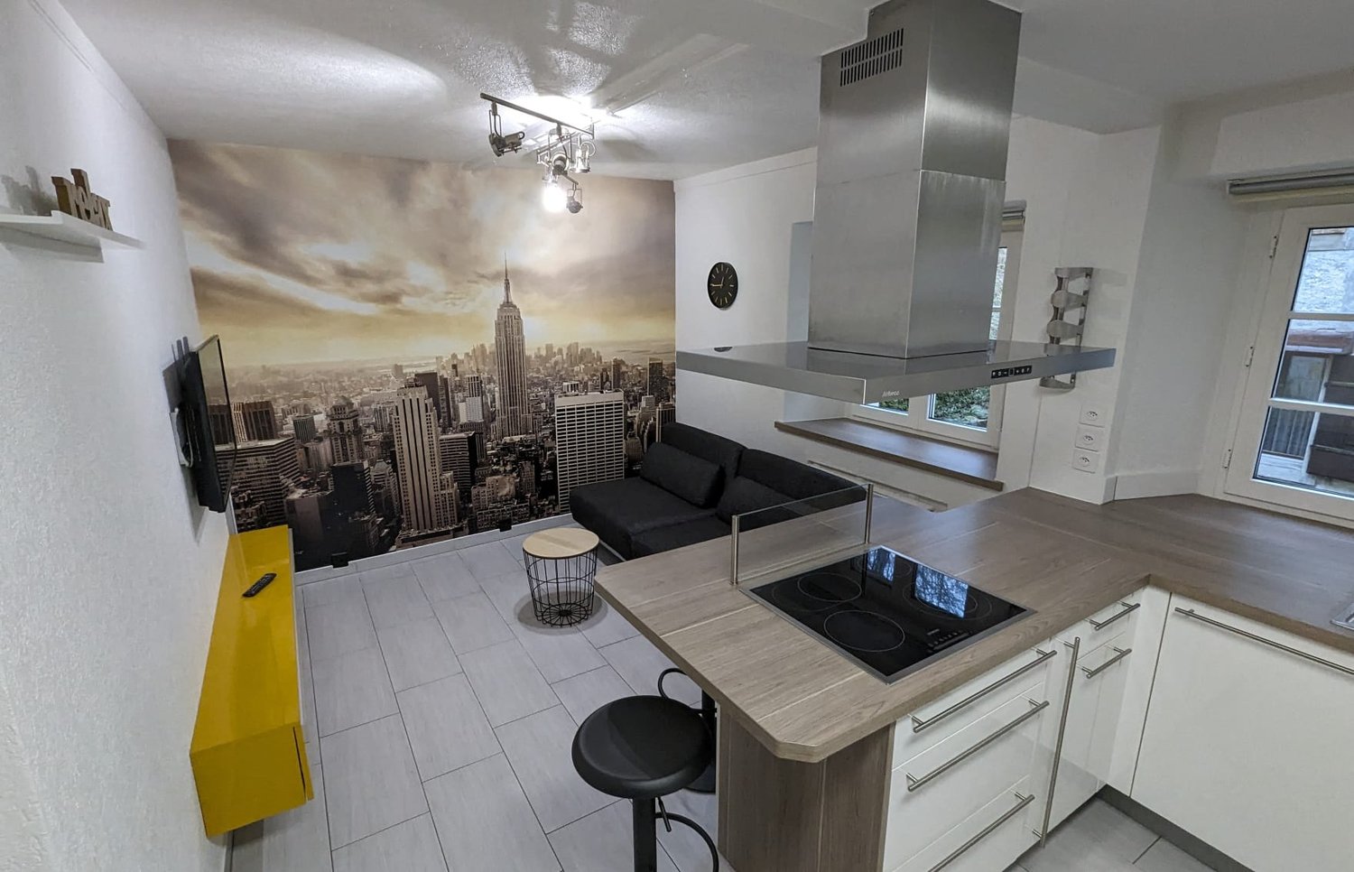 modern living room with kitchen and couch, modern kitchen with oven and stove, yellow wall art with remote control, large cityscape mural, white tiled floor