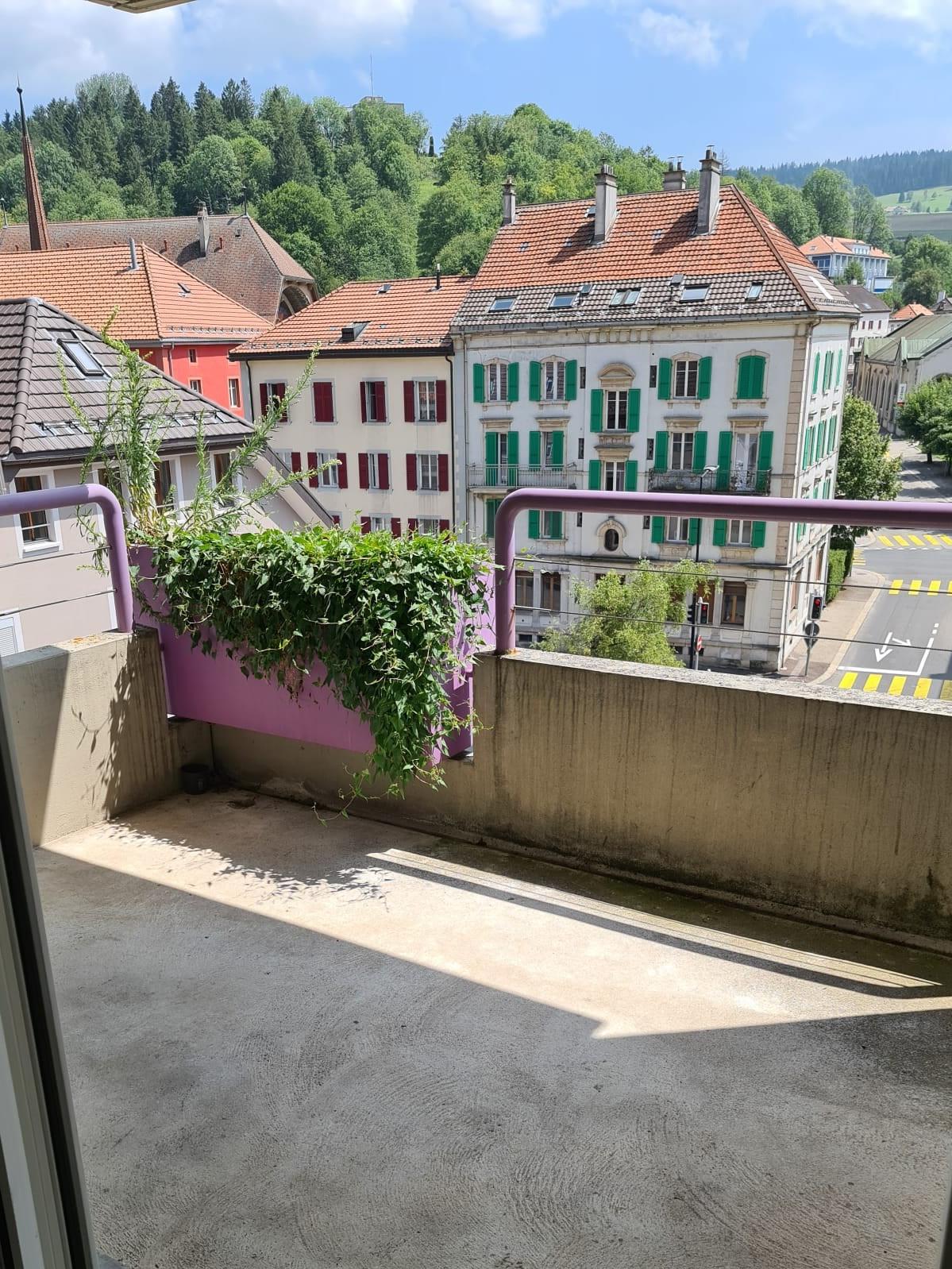 Empty balcony with concrete floor and railing, overlooking a street with trees, houses and hills.