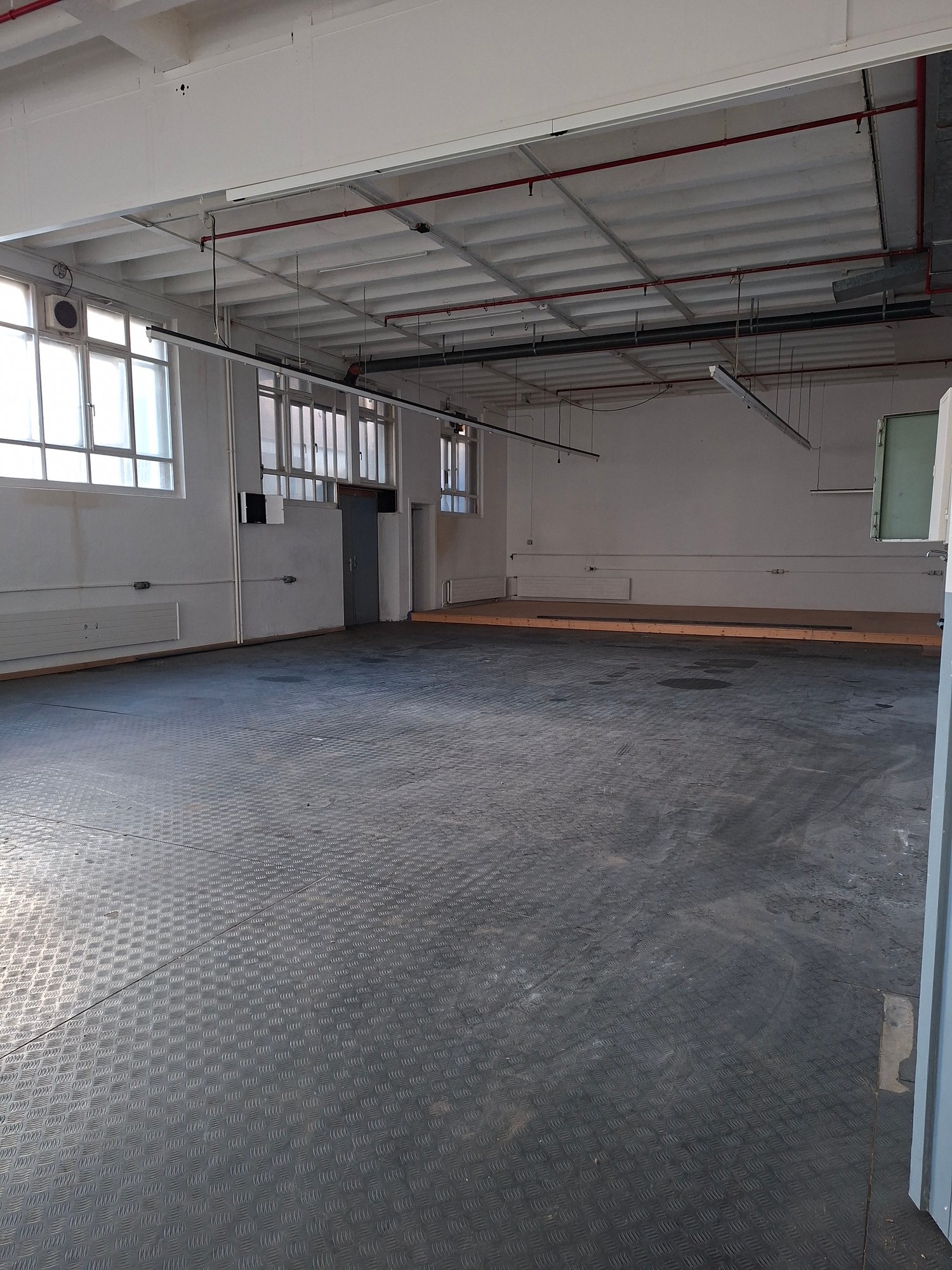 Large empty warehouse room, white walls, metal flooring, fluorescent ceiling lights, windows, radiators, small raised platform, door