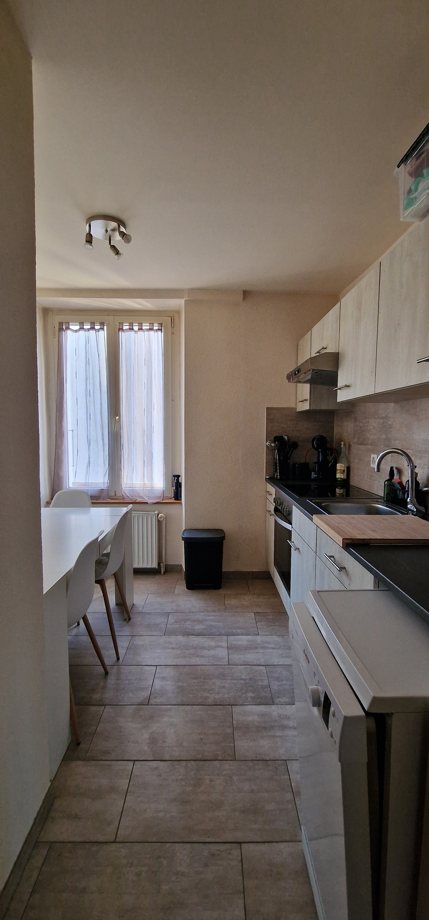 kitchen with dining area, white walls, wooden floor, white cabinets, white dining table, white chairs, white washing machine, dishwasher, white radiator, windows with white curtains, trash bin, ceiling lights