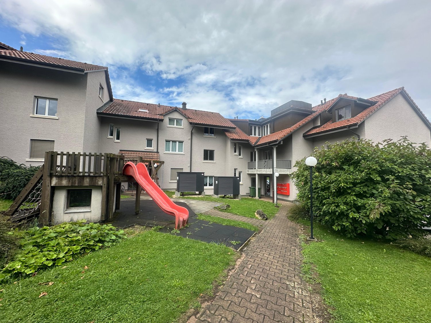 Multiple residential buildings, children's slide, lamp, paved pathway, well-kept grassy areas, trash bin, chimneys